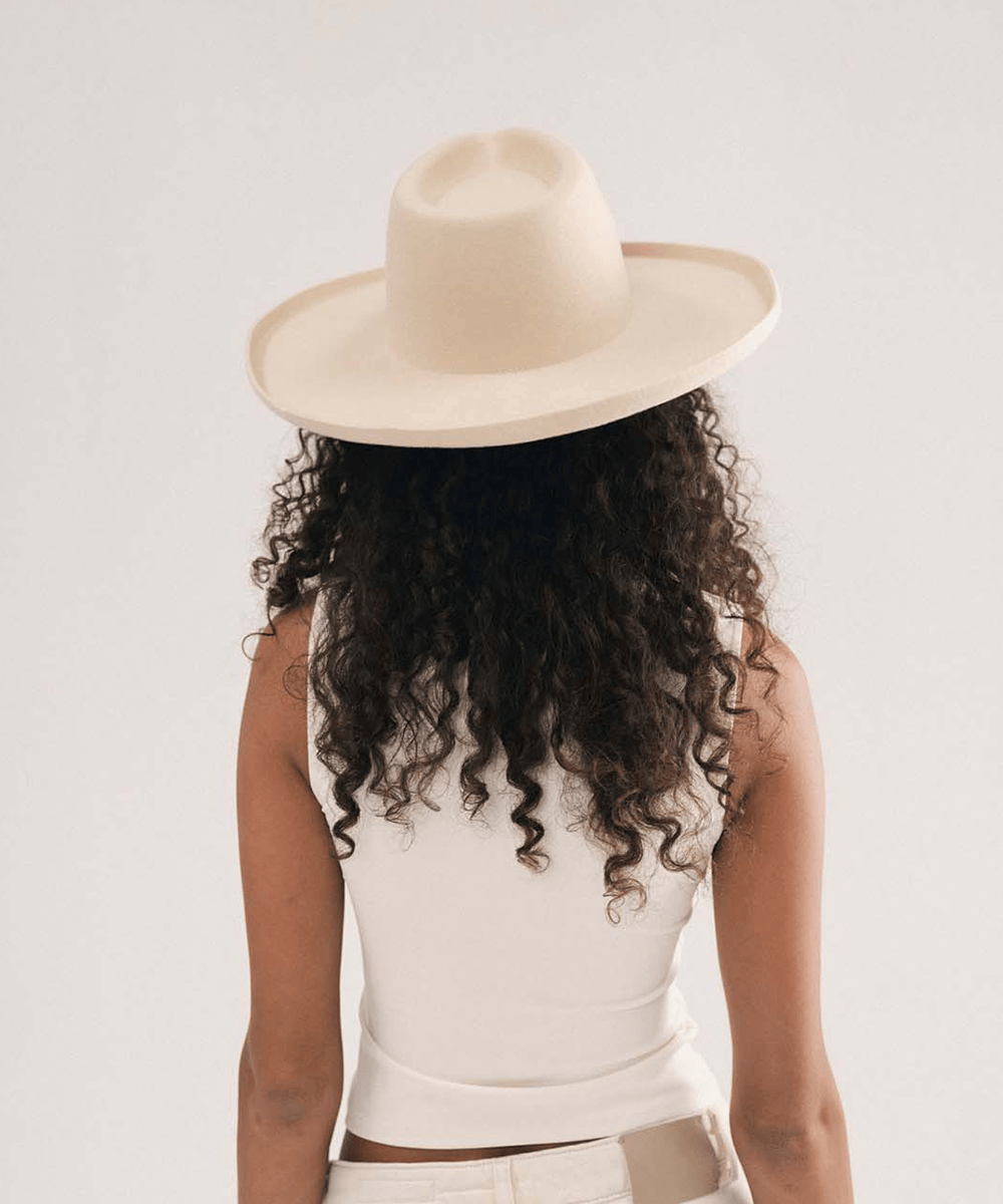 A person shown from behind wearing a wide-brim fedora hat, a white sleeveless top, and white pants against a plain light gray studio background #color_cream