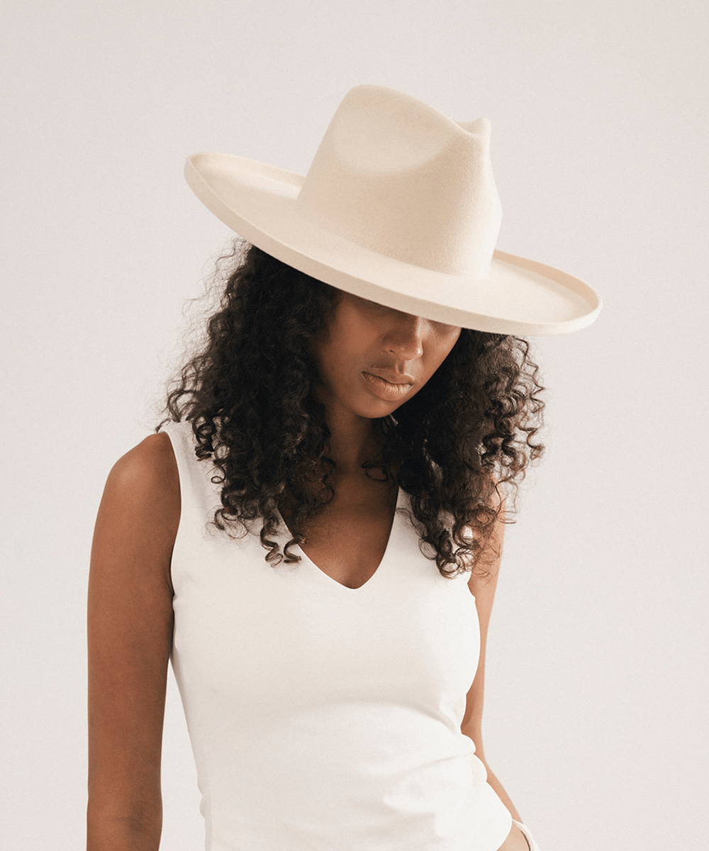A woman wearing a cream-colored wide-brim fedora hat and a matching cream sleeveless top, photographed against a plain light background #color_cream