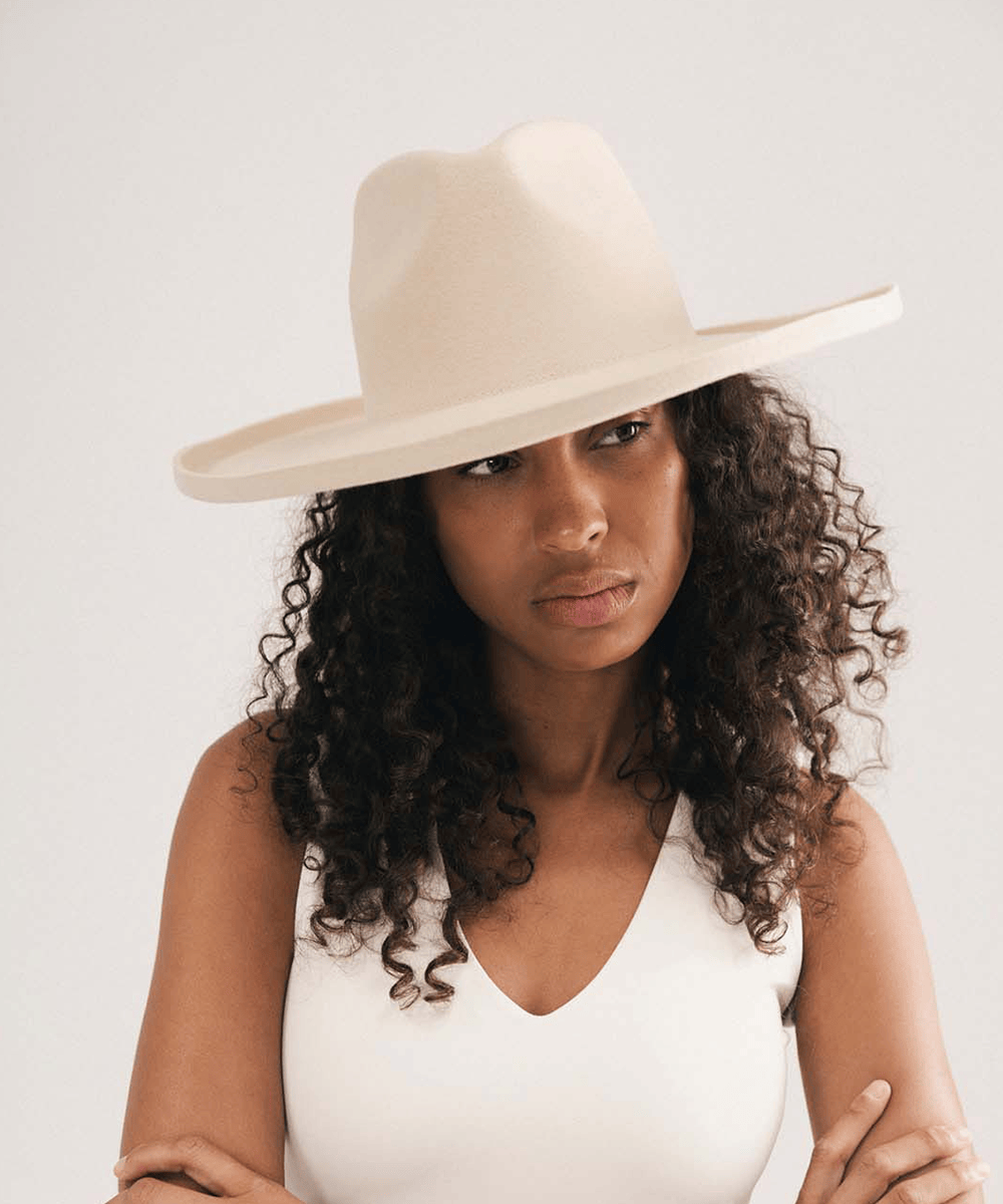 A woman with curly dark hair wearing a wide-brimmed cream-colored hat and a white sleeveless top, photographed against a plain light background #color_cream