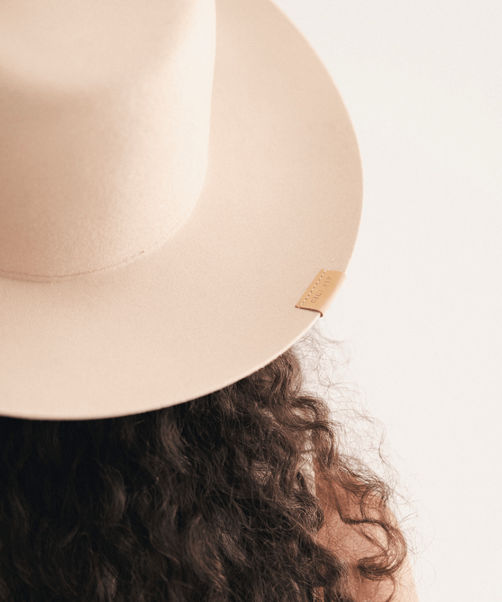 Close-up of a person wearing a wide-brimmed felt hat in a light beige color, viewed from behind, with dark curly hair visible against a plain white background #color_cream