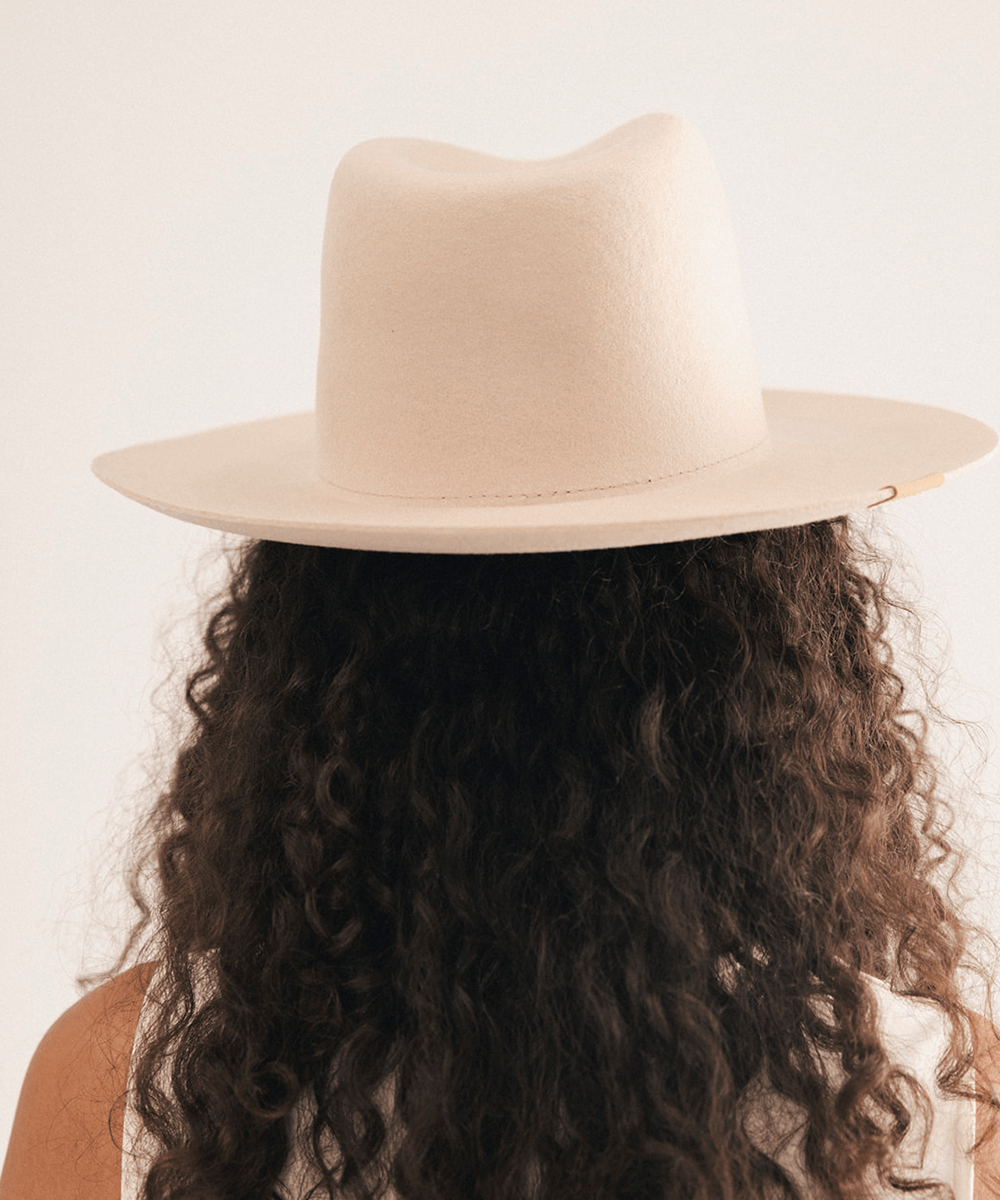 A person shown from the back wearing a wide-brim fedora hat, with long dark curly hair visible against a plain light background #color_cream