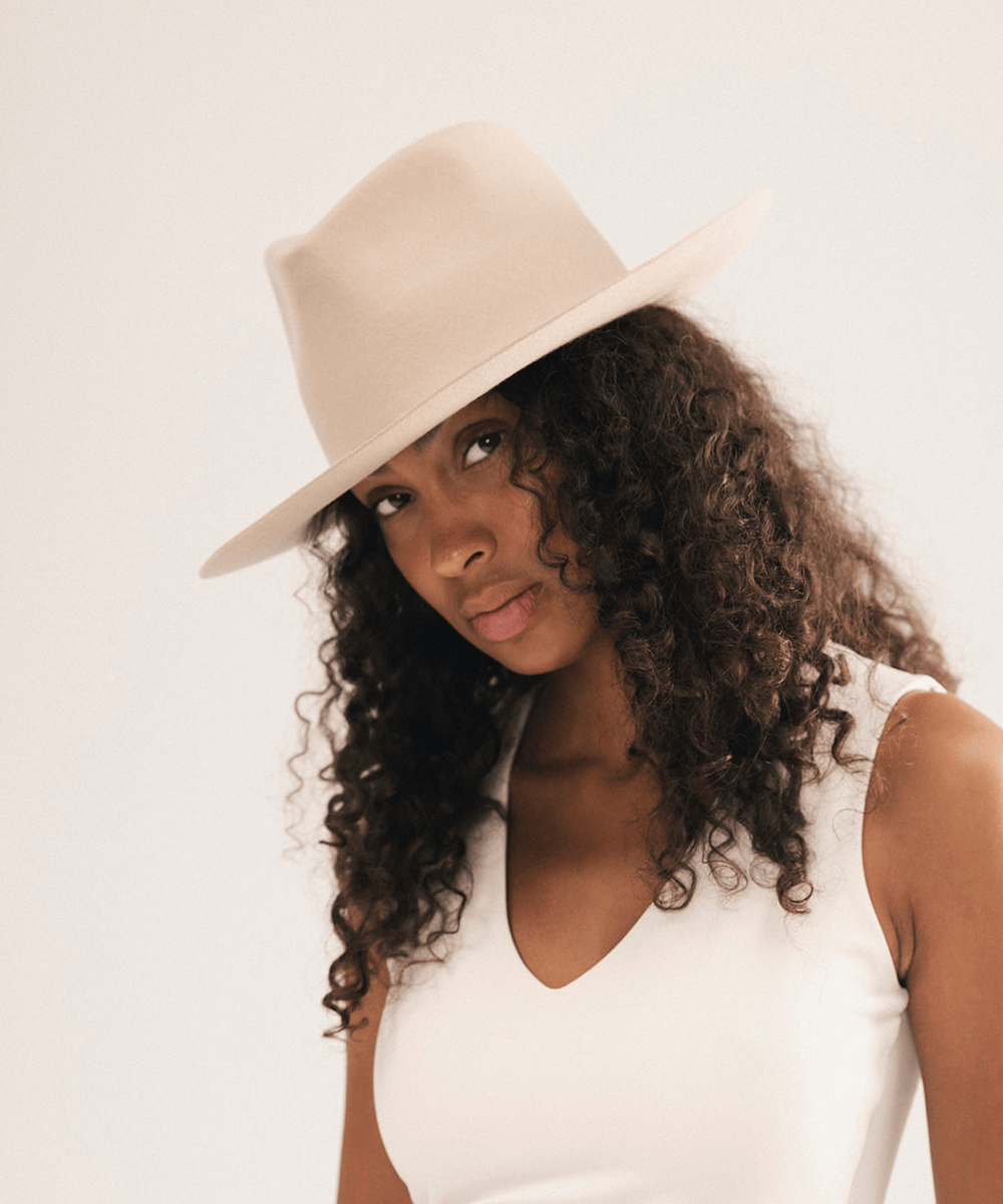 A woman with long, curly dark brown hair wearing a light beige wide-brim fedora hat and a white sleeveless top, photographed against a clean off-white studio background #color_cream