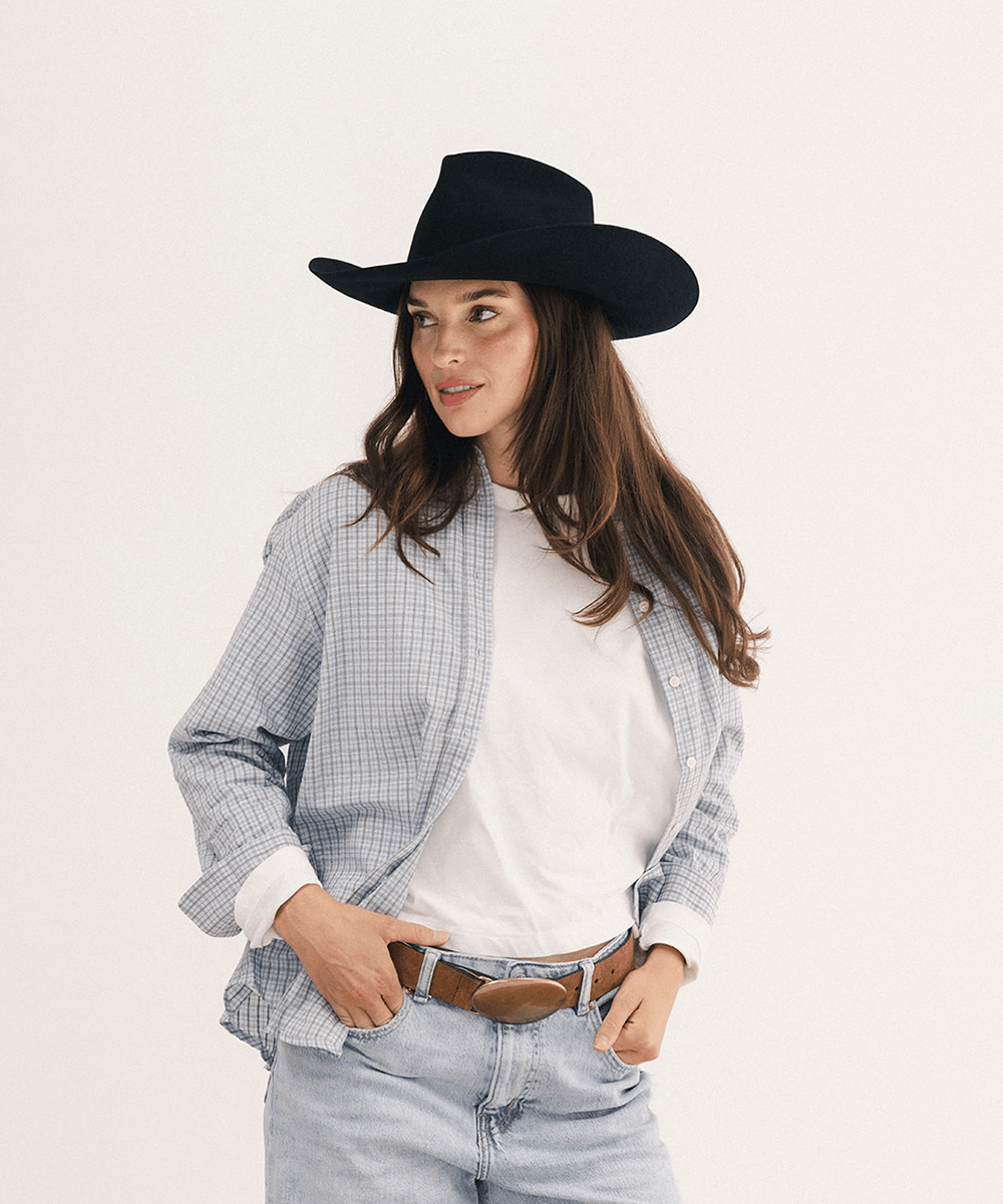 A woman wearing a black cowboy hat, a light blue and white gingham overshirt, a white t-shirt, and light-wash jeans with a brown belt, posing against a solid off-white background #color_black