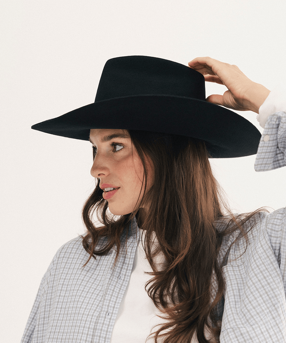 A woman wearing a wide-brim black cowboy hat and a light blue and white checkered shirt, shown in profile against a plain white background #color_black