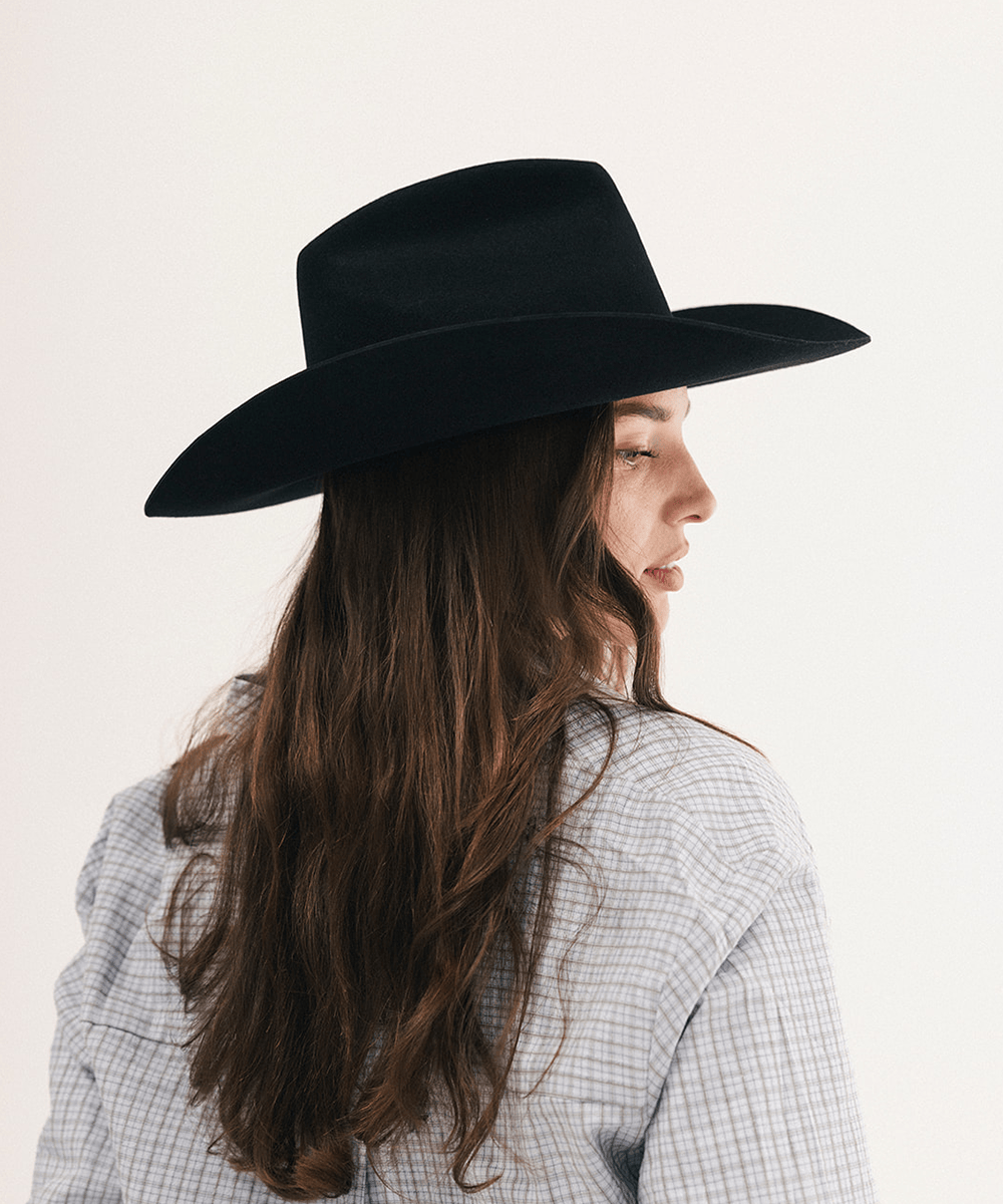 A woman with long brown hair is shown in profile wearing a black wide-brim fedora hat and a light blue and white checkered shirt against a plain off-white background #color_black