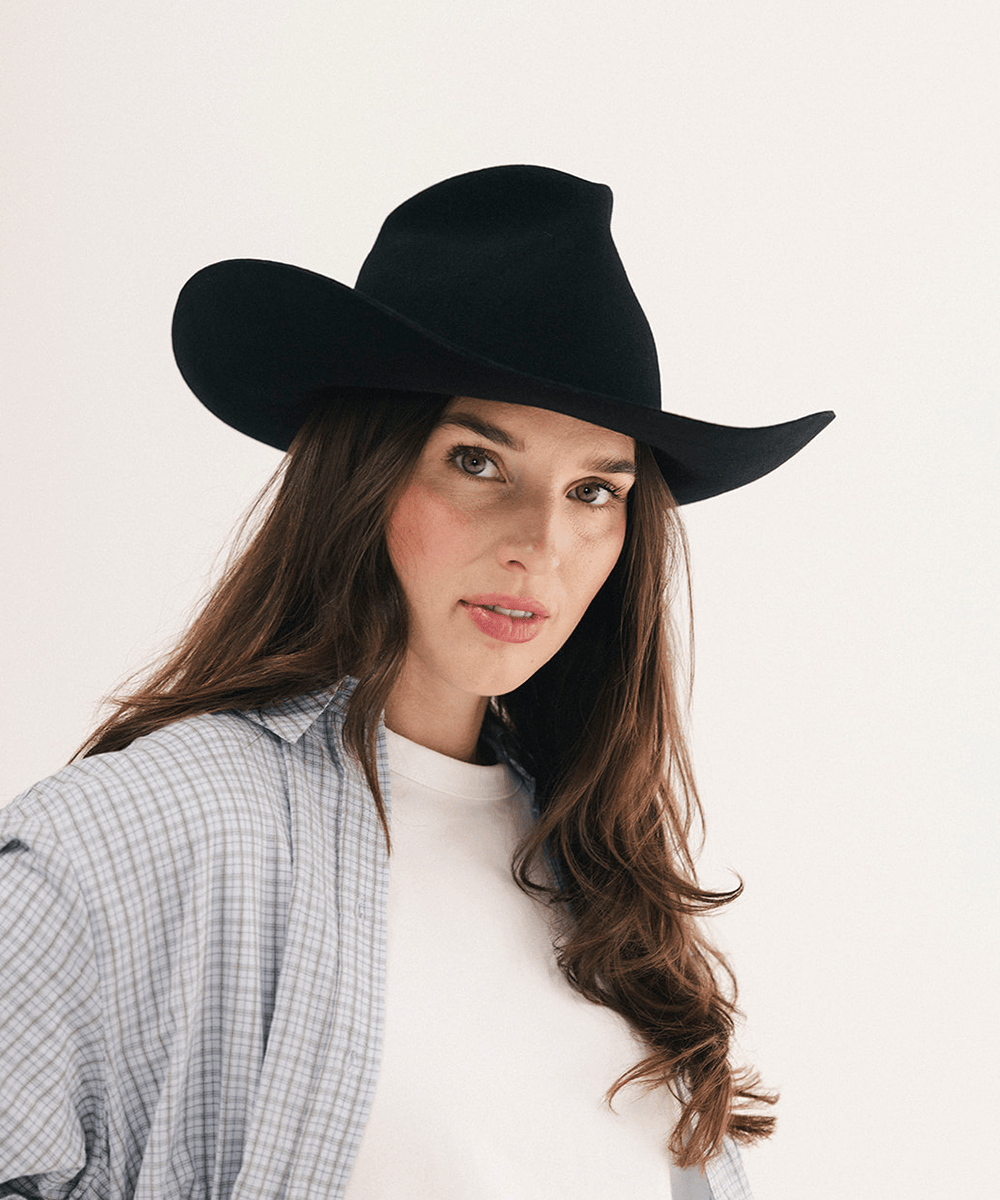 A woman wearing a black felt cowboy hat, a light blue and white plaid button-up shirt, and a white t-shirt, photographed against a clean off-white background #color_black