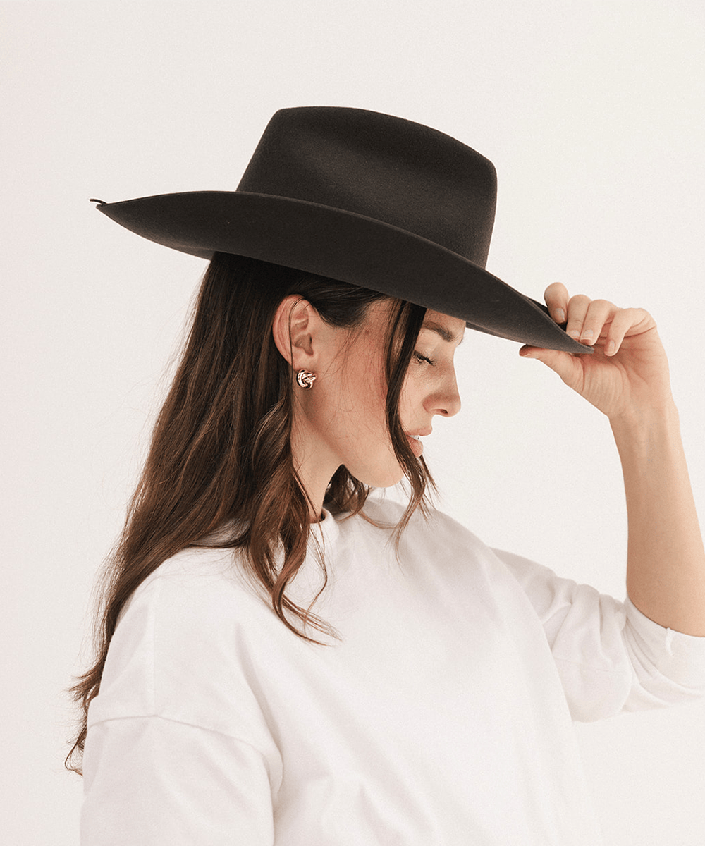 A woman wearing a black wide-brim fedora hat and a white long-sleeve shirt, shown in profile against a plain white studio background #color_dark brown