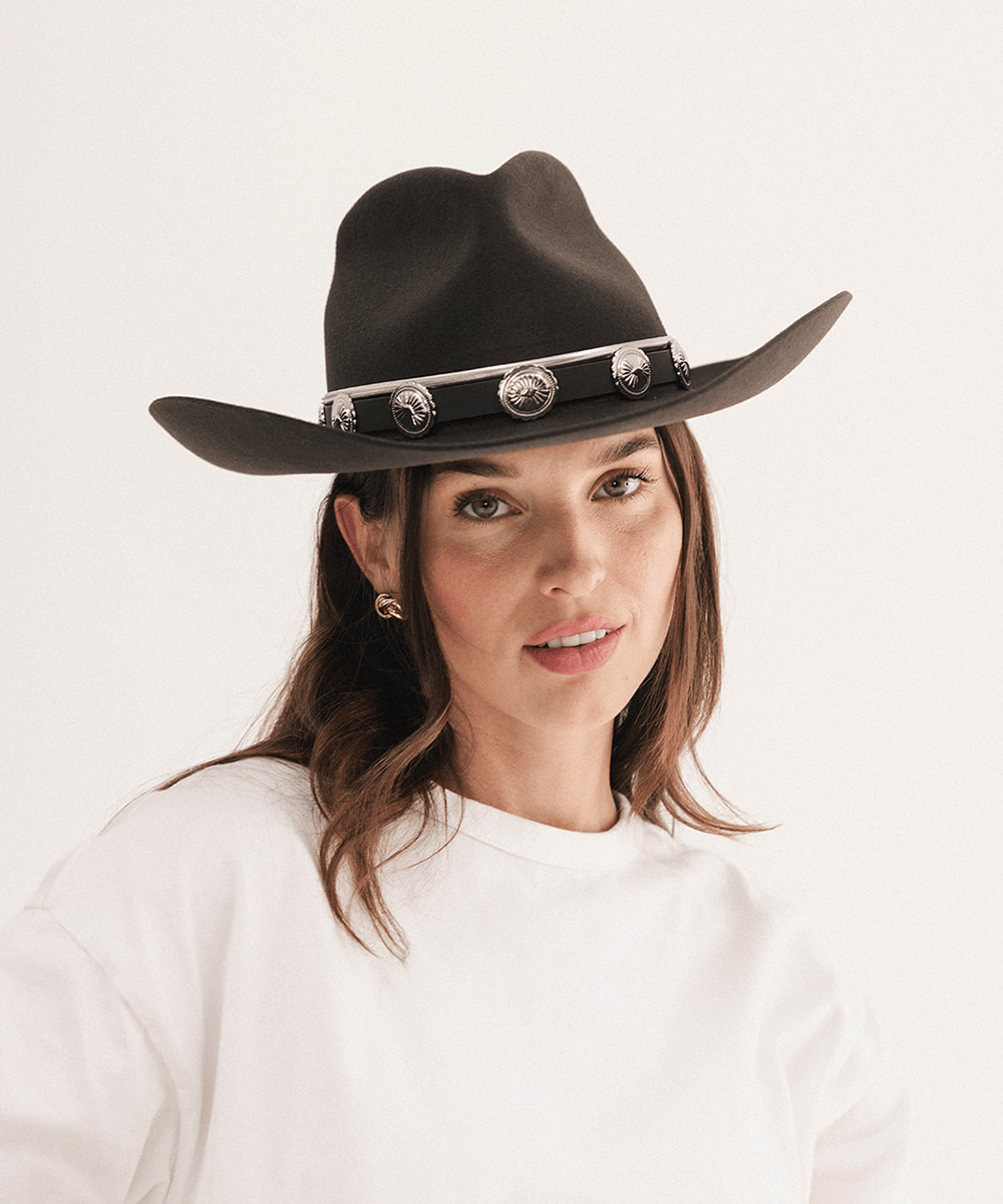 A woman wearing a dark brown felt cowboy hat with a black band and silver conchos, paired with a white crewneck sweatshirt, against a plain white background #color_dark brown