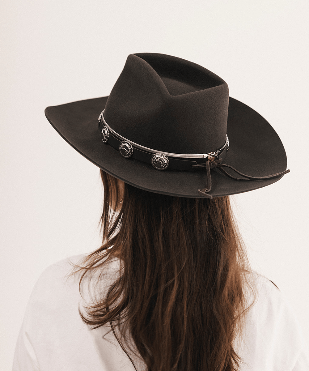 A woman viewed from behind wearing a dark brown felt cowboy hat with a silver concho strap, against a plain off-white background #color_dark brown