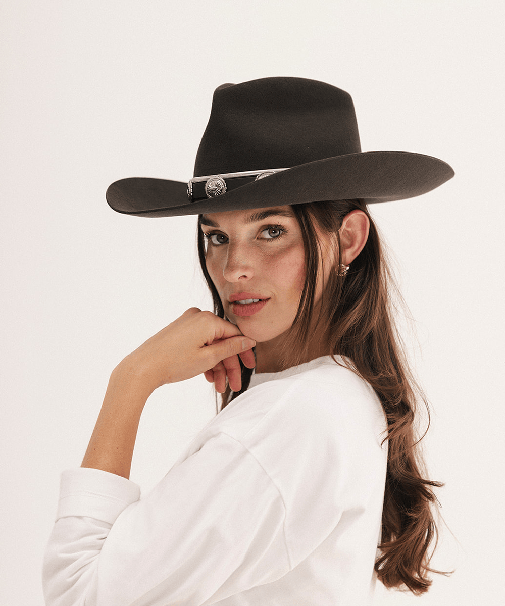 A woman wearing a black wide-brim fedora hat with a silver decorative band and a white long-sleeve shirt, photographed against a clean off-white studio background #color_dark brown