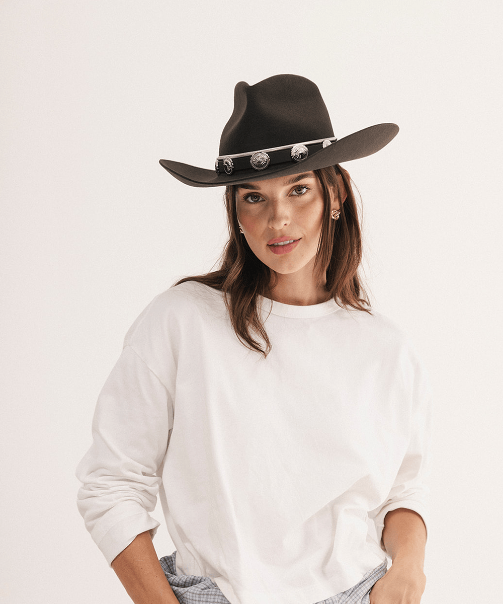 A woman wearing a dark brown felt cowboy hat with silver conchos and a white long-sleeve shirt, photographed against a clean off-white studio background #color_dark brown