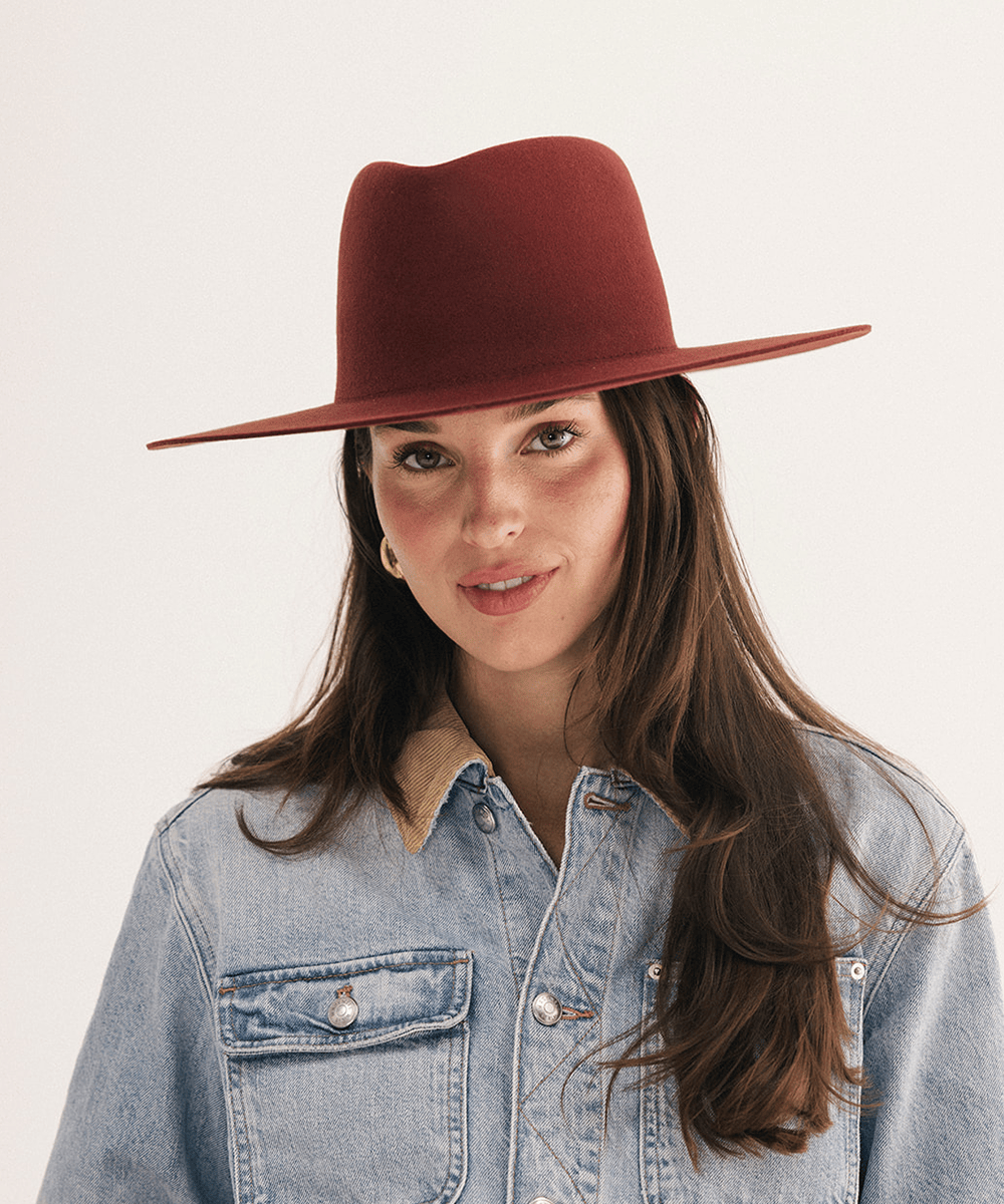 A woman wearing a wide-brimmed felt hat in a deep burgundy red, paired with a light blue denim shirt. She is photographed against a clean, off-white studio background #color_rusty red