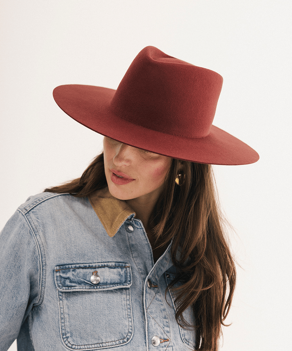 A woman wearing a wide-brimmed felt hat in a muted terracotta red, paired with a light blue denim jacket and gold hoop earrings, photographed against a clean white background #color_rusty red