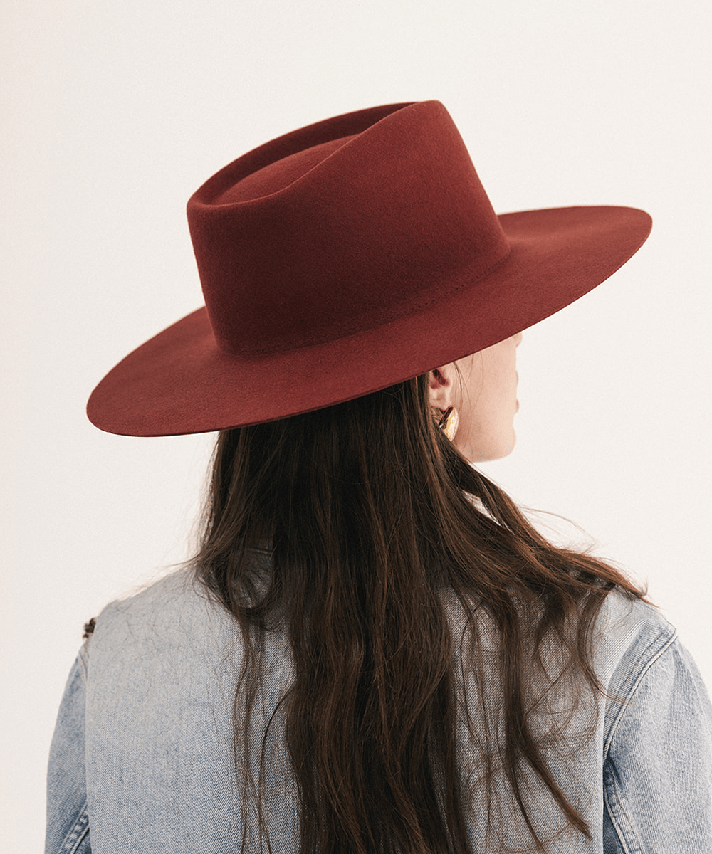 A woman shown from the back wearing a wide-brimmed felt hat in a deep burgundy red, paired with a light blue denim jacket and gold hoop earrings, against a plain off-white background #color_rusty red