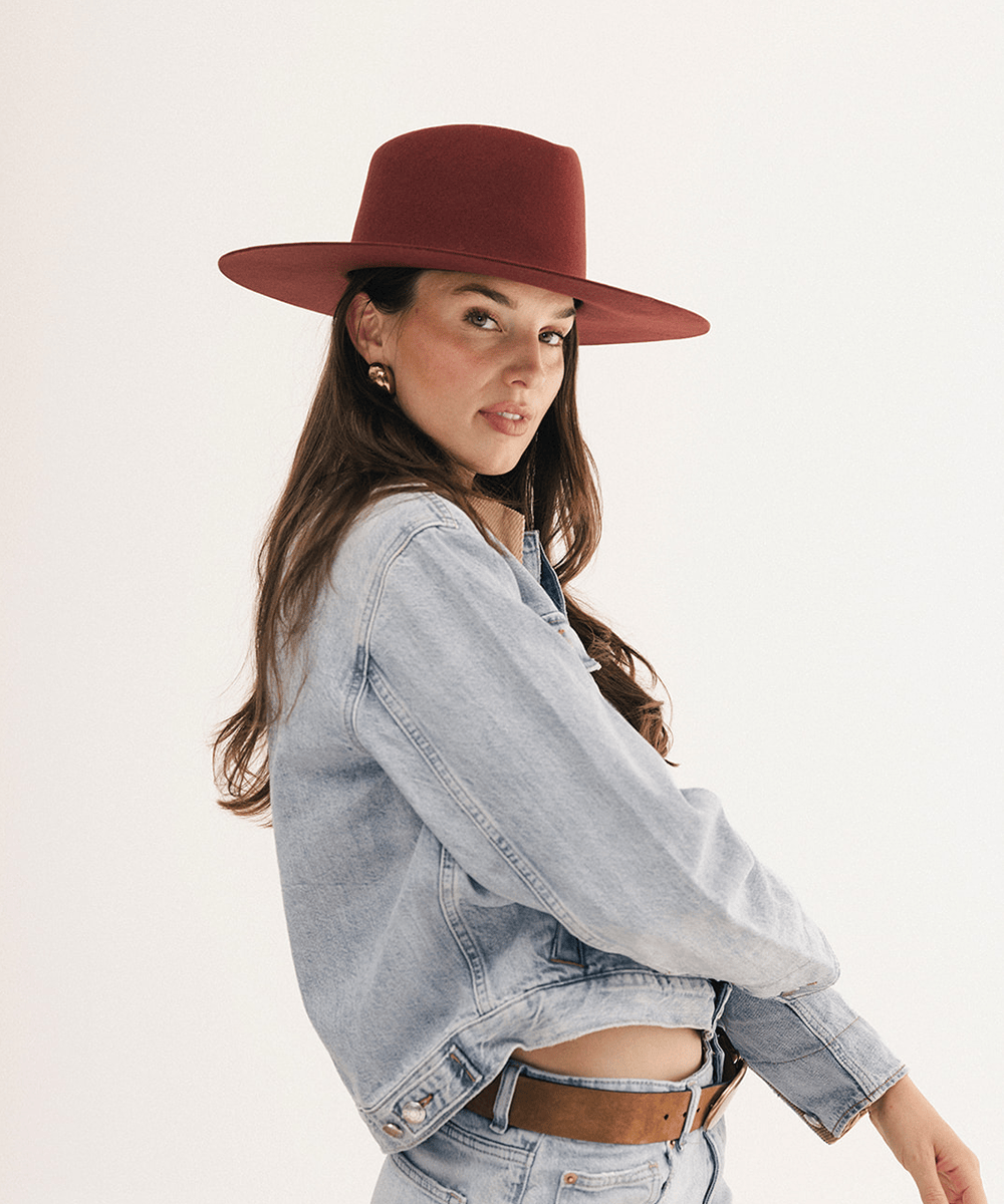 A woman wearing a wide-brimmed rust-brown felt hat, a light-wash denim jacket, and a brown belt, photographed against a clean off-white studio background #color_rusty red