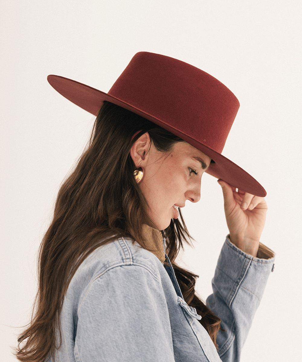 A woman in profile wearing a wide-brimmed felt hat in a deep burgundy red, styled with a light blue denim jacket and gold earrings against a plain white background #color_rusty red