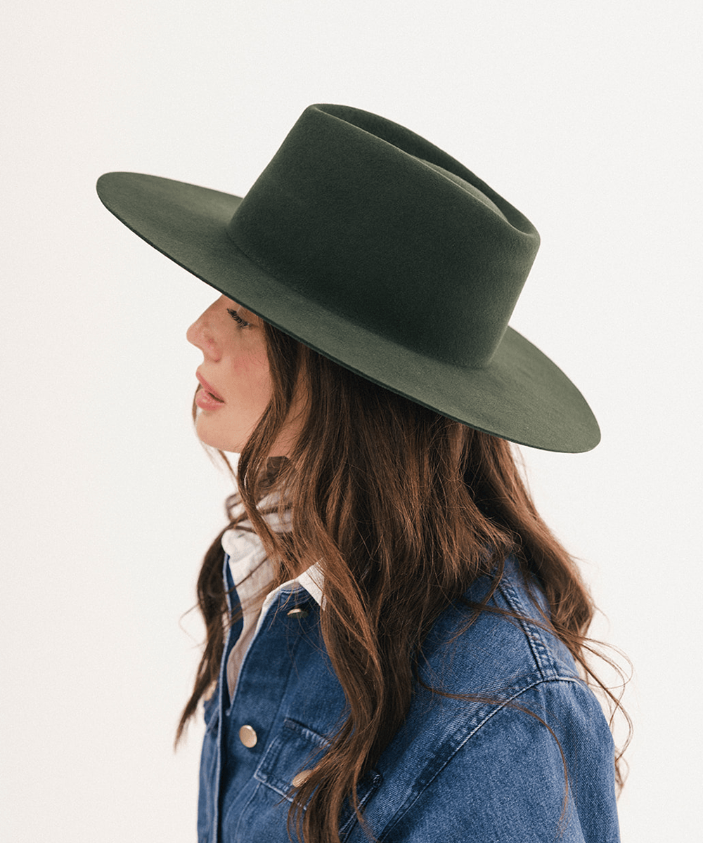 A woman in profile wearing a dark olive green wide-brim fedora hat and a blue denim jacket, photographed against a clean white studio background #color_dark green