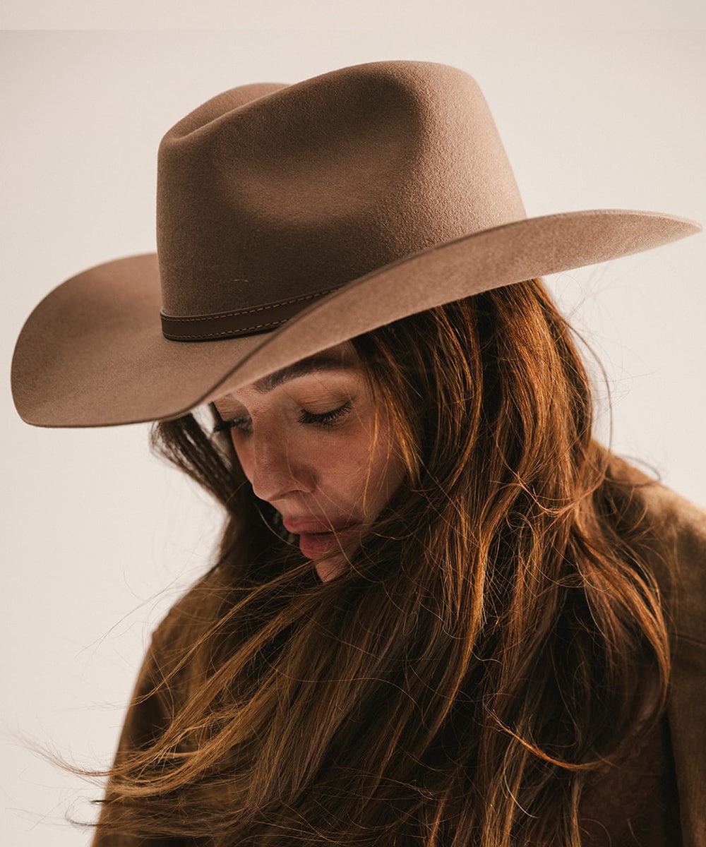 Woman wearing a chocolate colored cowboy hat with a hat band and a brown jacket, looking down against a plain background #color_chocolate