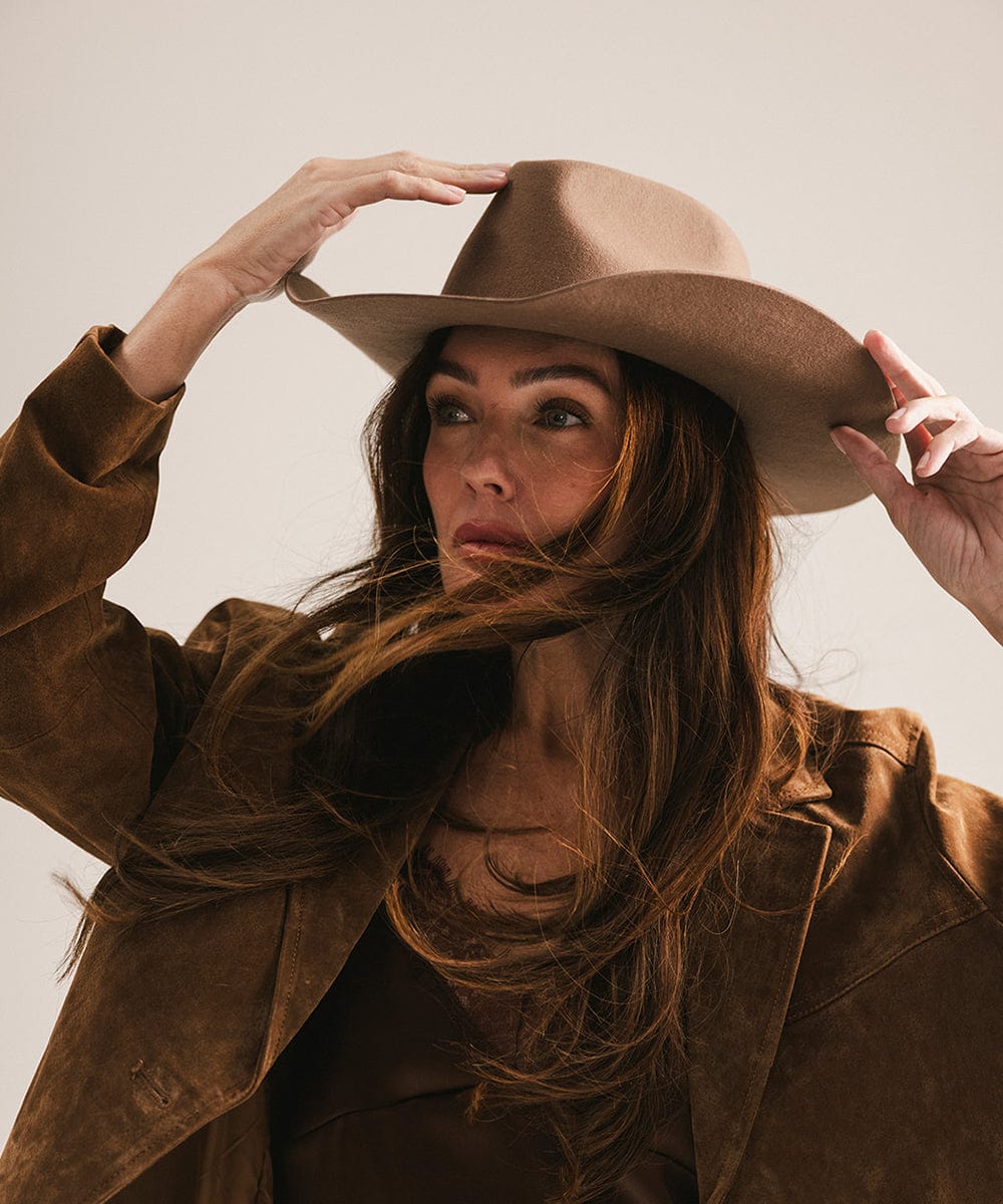 Woman wearing a chocolate colored cowboy hat with a hat band and a brown top with a brown jacket against a plain background #color_chocolate