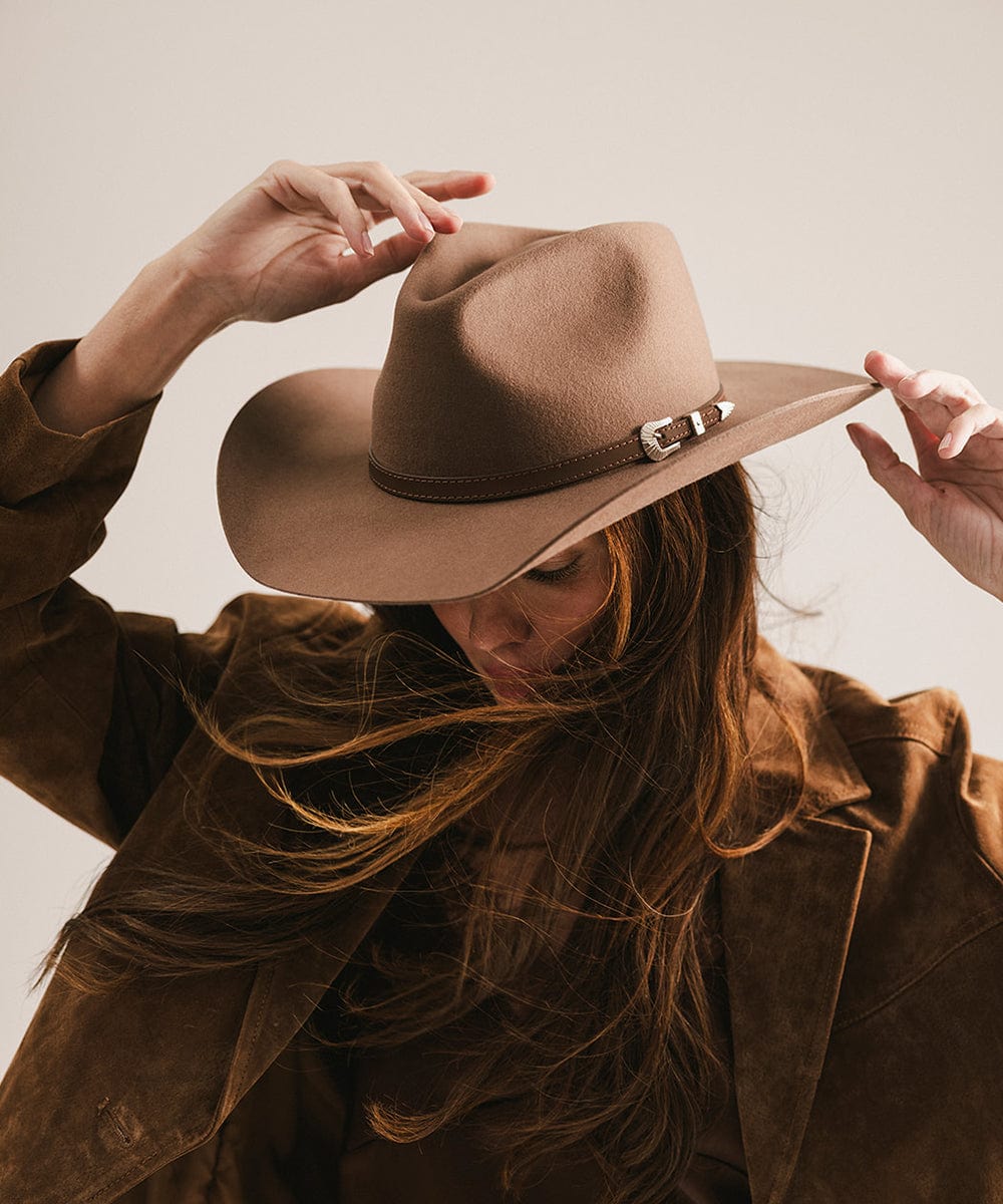 Woman wearing a chocolate colored cowboy hat with a hat band and a brown top with a brown jacket, looking down against a plain background #color_chocolate