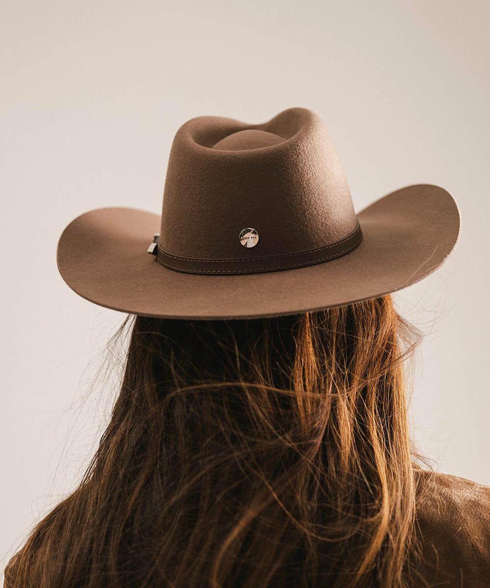 Woman wearing a chocolate colored cowboy hat with a hat band and a brown jacket, facing behind against a plain background #color_chocolate