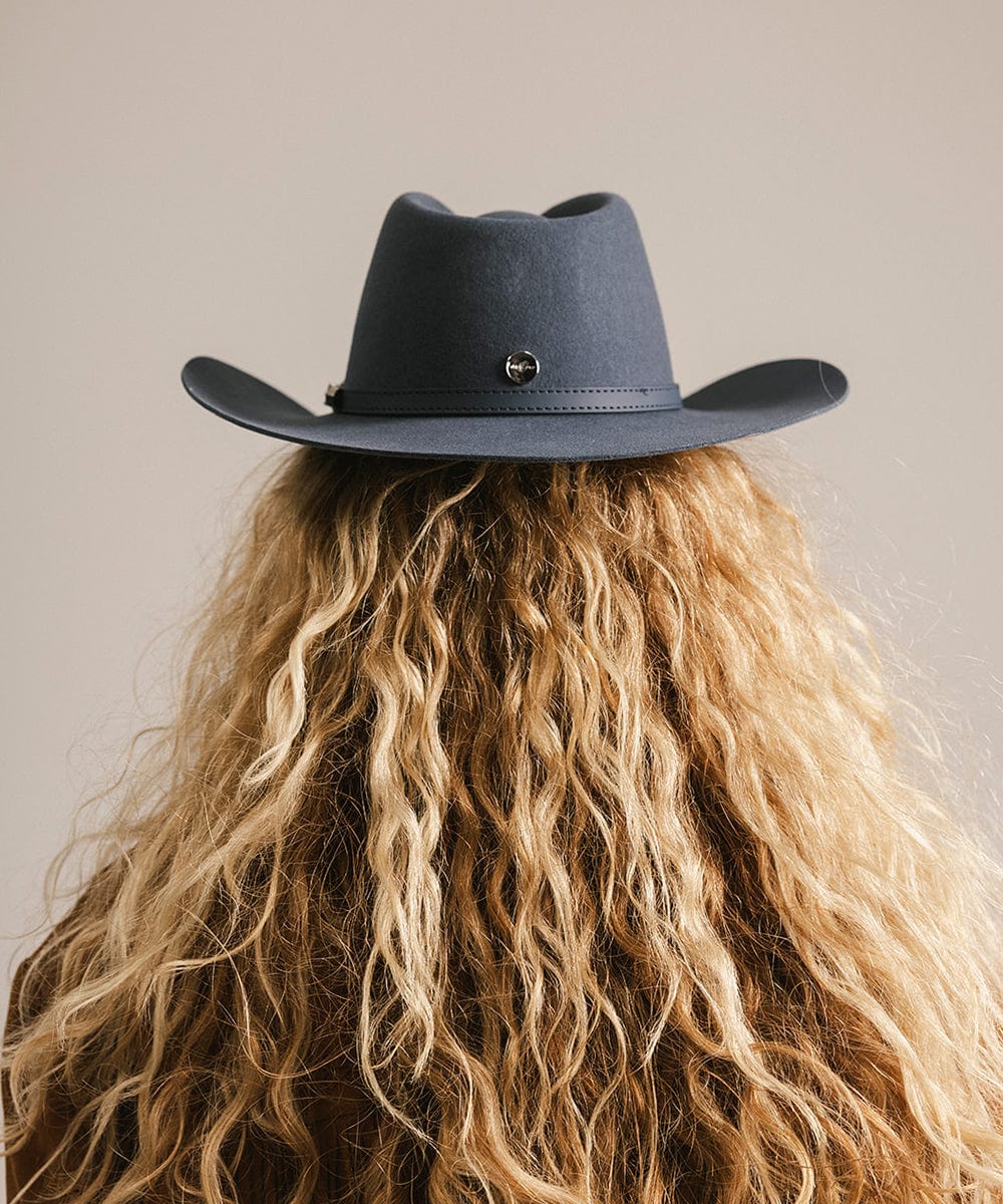 Woman wearing a vintage blue colored cowboy hat with a hat band  and a brown jacket, facing behind against a plain background #color_vintage blue