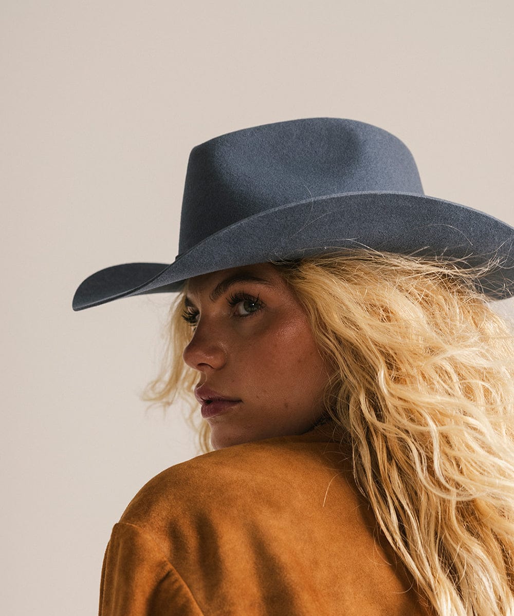 Woman wearing a vintage blue colored cowboy hat with a hat band  and a brown jacket, looking sideways against a plain background #color_vintage blue