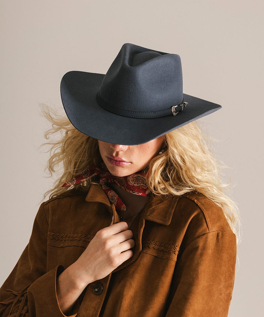 Woman wearing a vintage blue colored cowboy hat with a hat band  and a brown jacket against a plain background #color_vintage blue