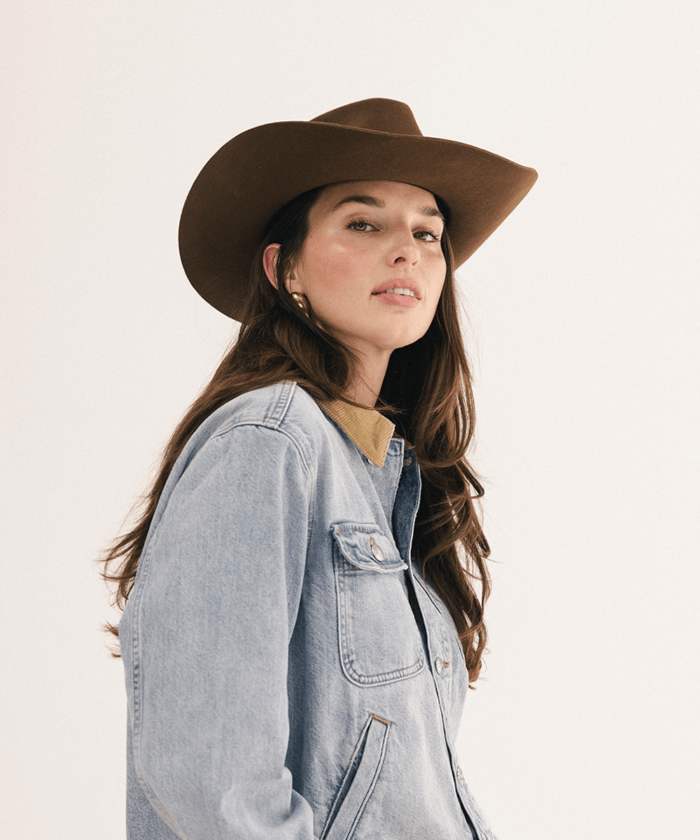 A woman wearing a brown wide-brim cowboy hat and a light-wash denim jacket over a tan top, photographed against a plain off-white studio background #color_coffee