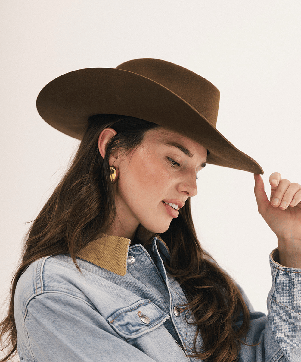 A woman wearing a brown wide-brim cowboy hat and a light blue denim jacket with a tan corduroy collar, posing against a plain off-white background #color_coffee