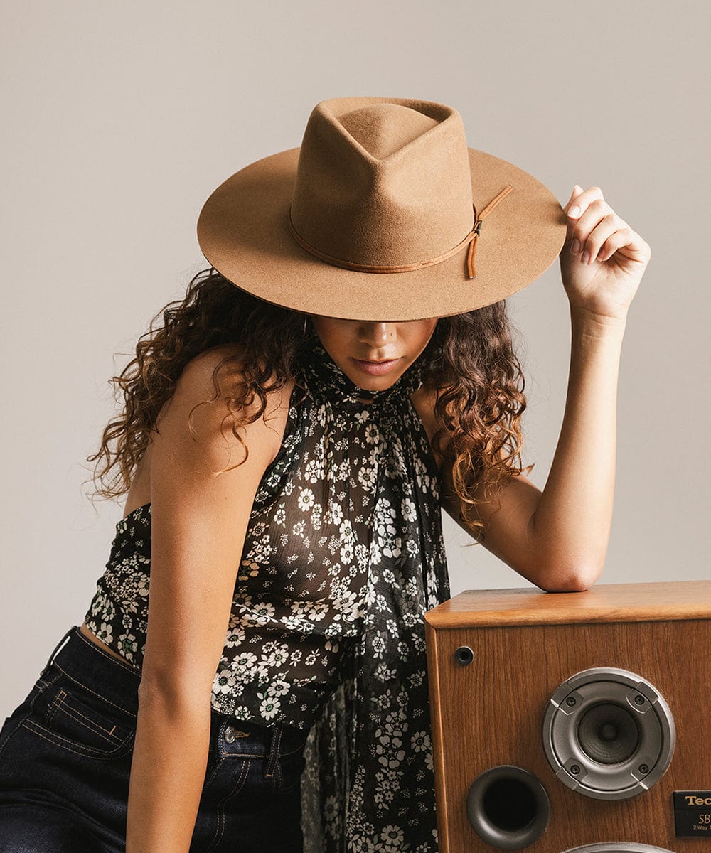 Woman wearing a caramel colored fedora hat with a hat band and a floral top, standing next to a wooden speaker against a plain background #color_caramel