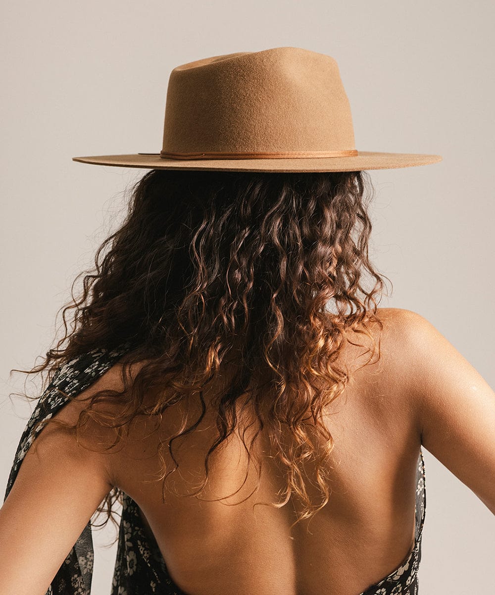 Woman wearing a caramel colored fedora hat with a hat band and a floral top, facing behind against a plain background #color_caramel