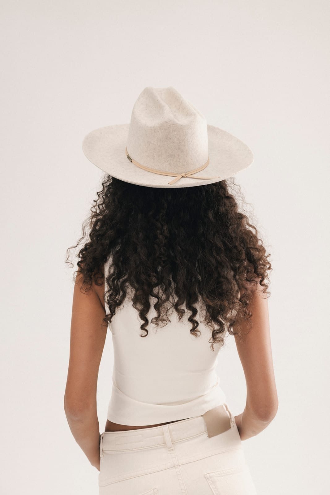 A woman with long, curly dark hair is shown from behind, wearing a light beige wide-brim fedora hat, a white sleeveless top, and white pants against a plain white studio background #color_mix grey
