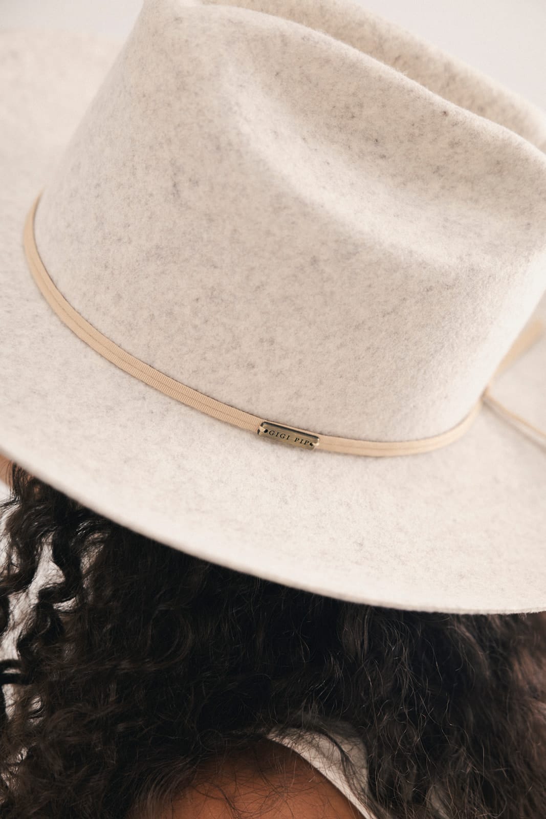 Close-up of a person wearing a wide-brim fedora hat in a light oatmeal beige felt, with a thin tan band and a small gold-tone metal logo plaque on the front #color_mix grey