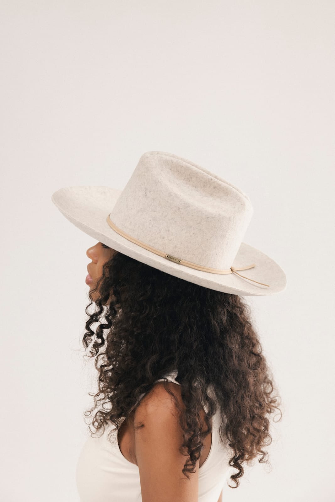 A woman in profile wearing a wide-brimmed felt hat in a light cream color with a thin tan band, set against a plain off-white background #color_mix grey