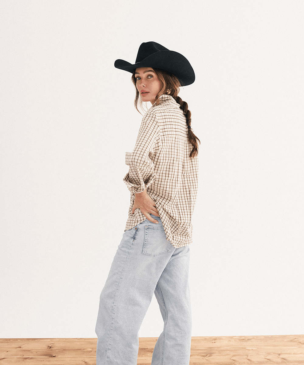 A woman wearing a black cowboy hat, a beige and brown checkered long-sleeve shirt, and light-wash blue jeans, standing against a plain white wall with a wooden floor #color_black
