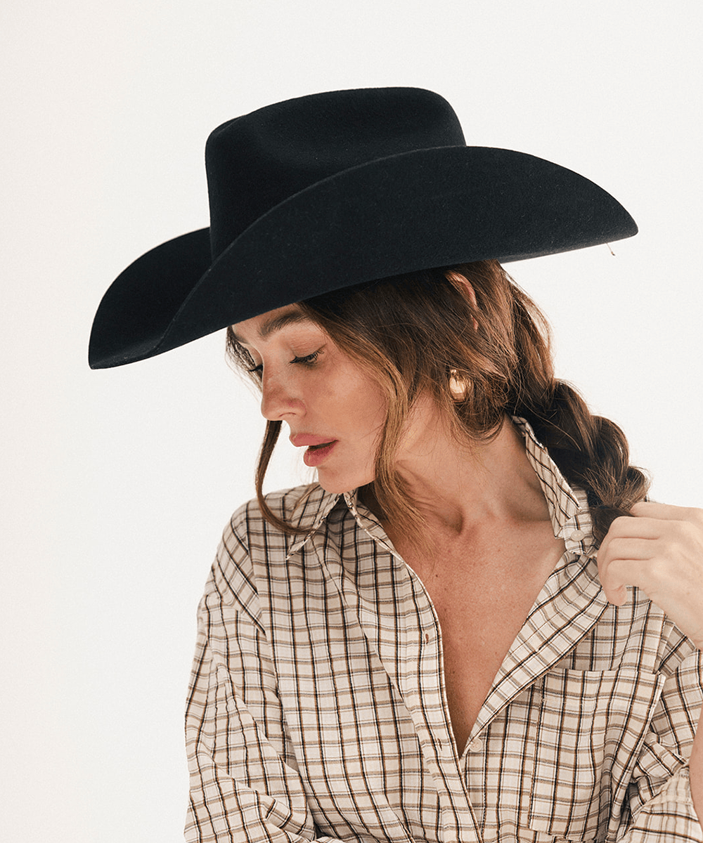 A woman wearing a black wide-brim cowboy hat and a beige and brown plaid button-down shirt, photographed against a clean white background #color_black