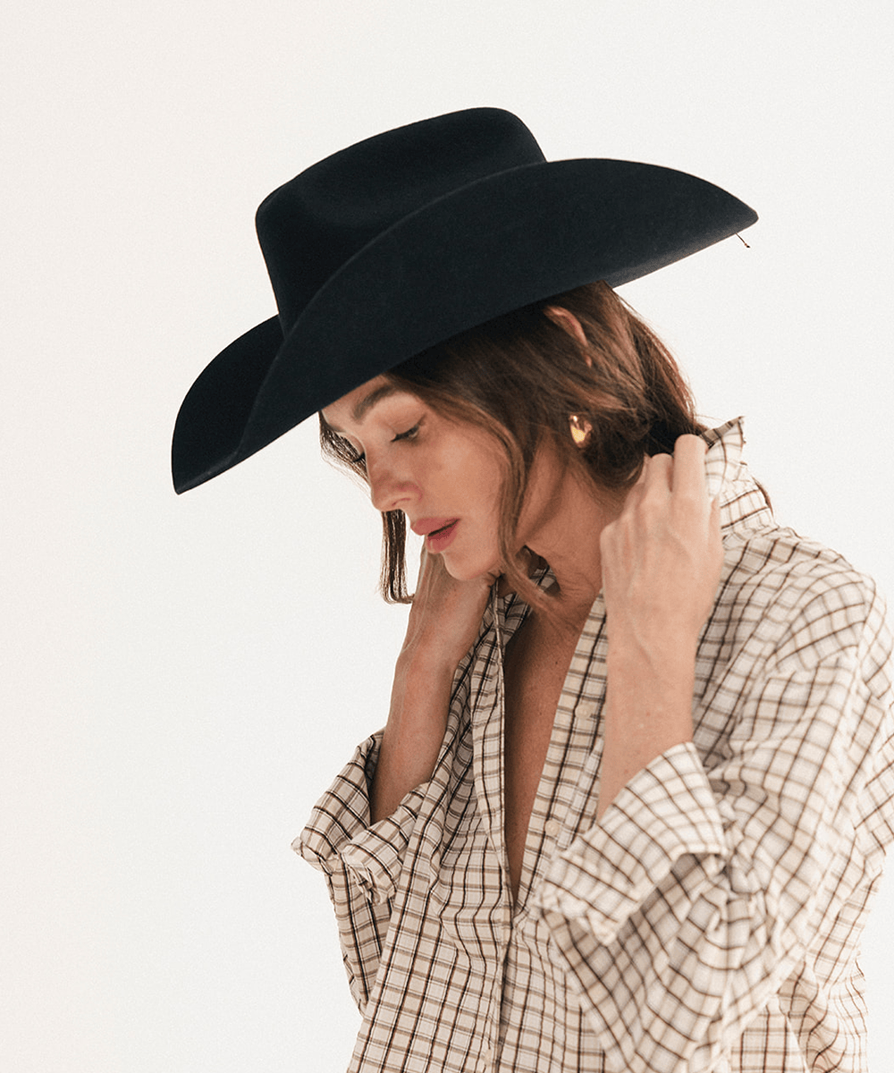 A woman wearing a black wide-brim fedora hat and a beige and brown plaid button-down shirt, posing against a solid off-white background #color_black