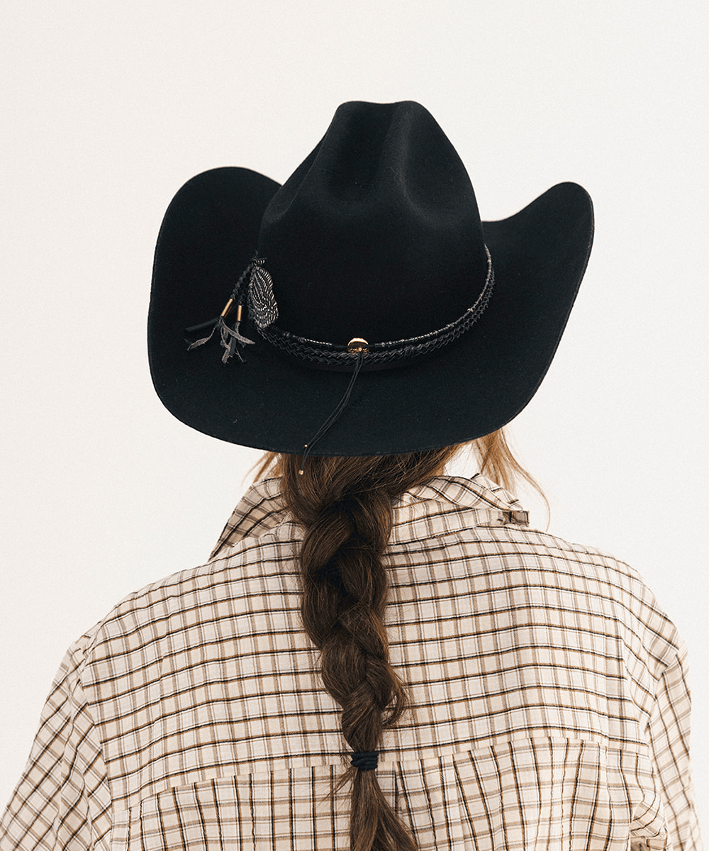 A person shown from behind wearing a black cowboy hat with a braided band and feather detail, a beige plaid shirt, and a long brown braid against a plain light background #color_black