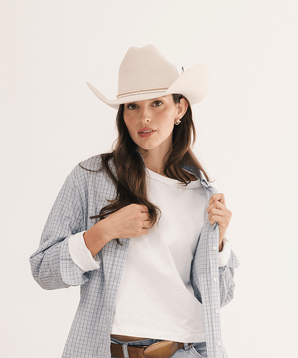 A woman modeling a light beige cowboy hat, a white crewneck t-shirt, and a light blue and white plaid button-up shirt over a white tee, set against a plain white studio background #color_off white