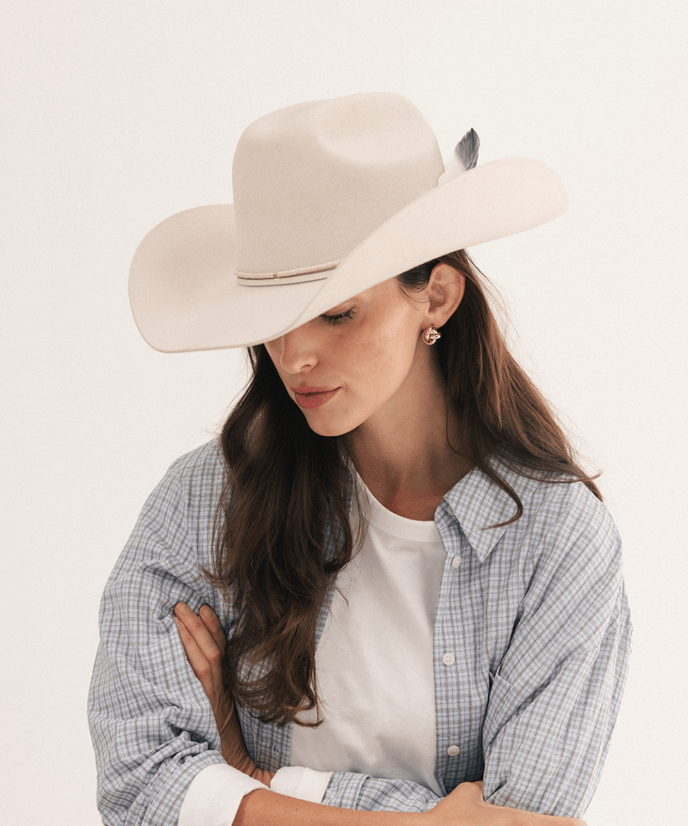 A woman wearing a cream-colored cowboy hat with a feather accent, a blue and white plaid button-down shirt, and a white t-shirt underneath, photographed against a clean white background #color_off white
