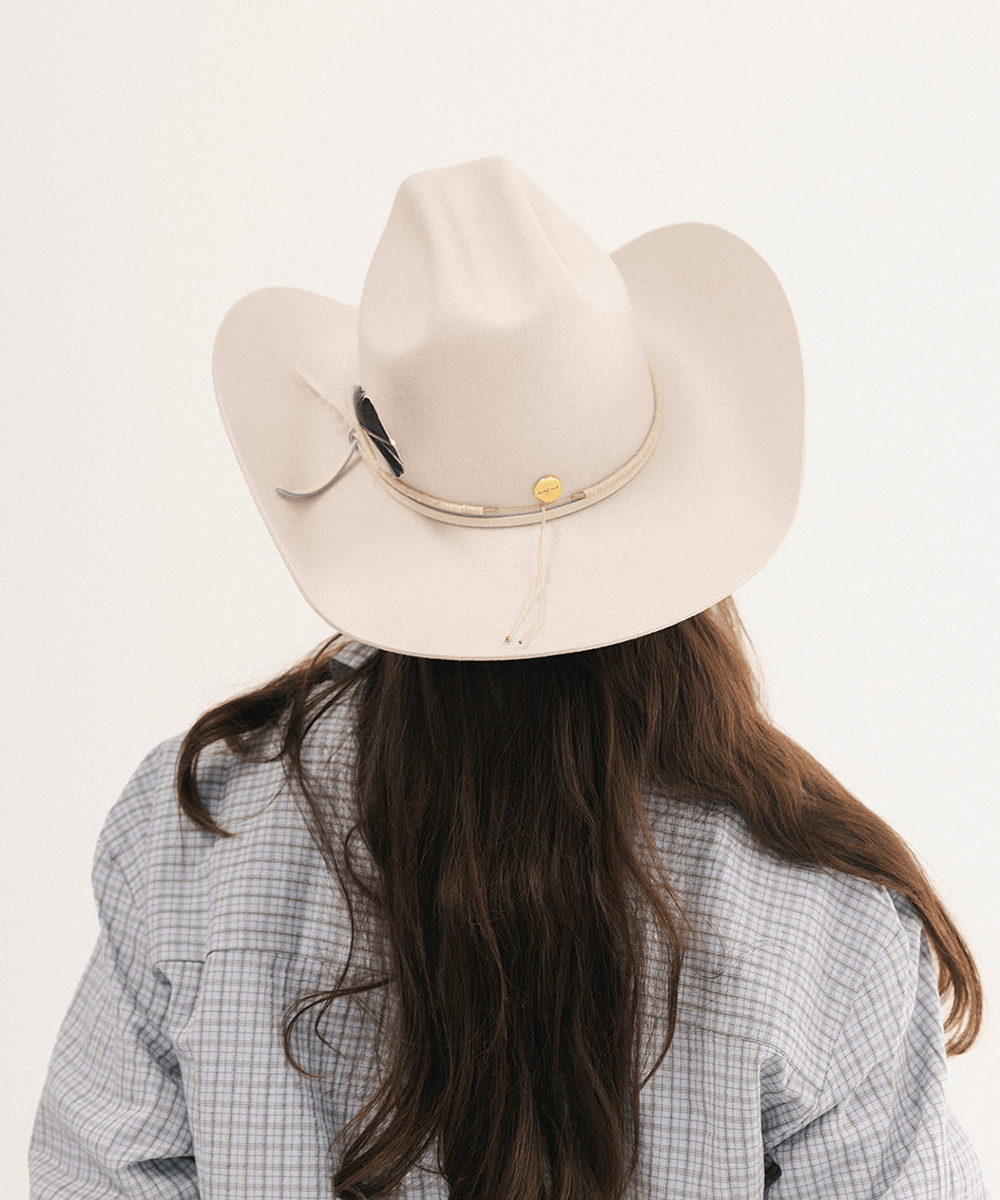 A person viewed from behind wearing a light cream-colored cowboy hat with a beige band, a small gold button, and a black feather accent, paired with a blue and white checkered shirt against a plain white background #color_off white