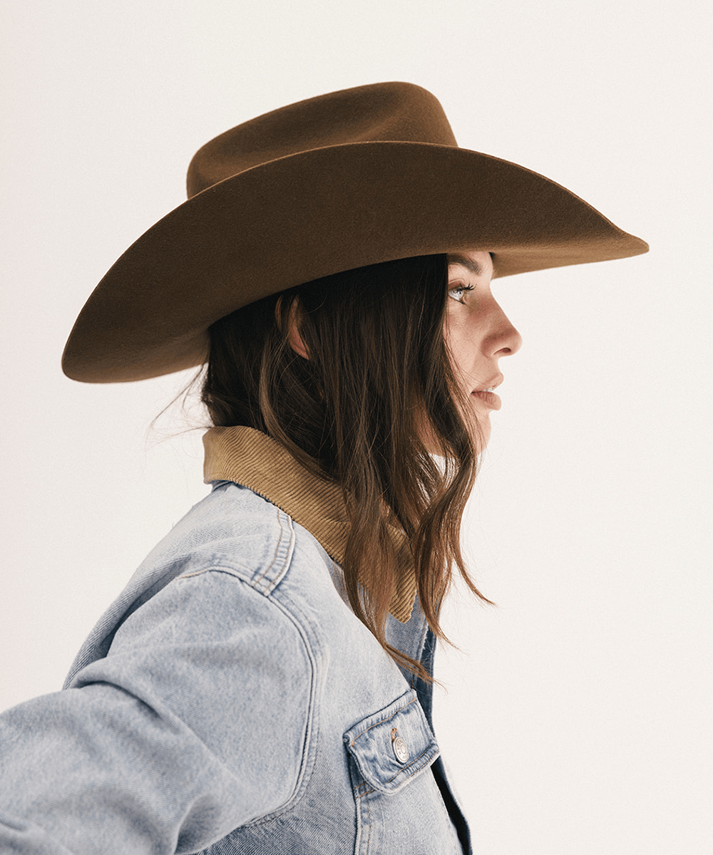 A woman in profile wearing a brown wide-brim fedora hat, a light blue denim jacket, and a tan corduroy collar, set against a plain off-white background #color_coffee