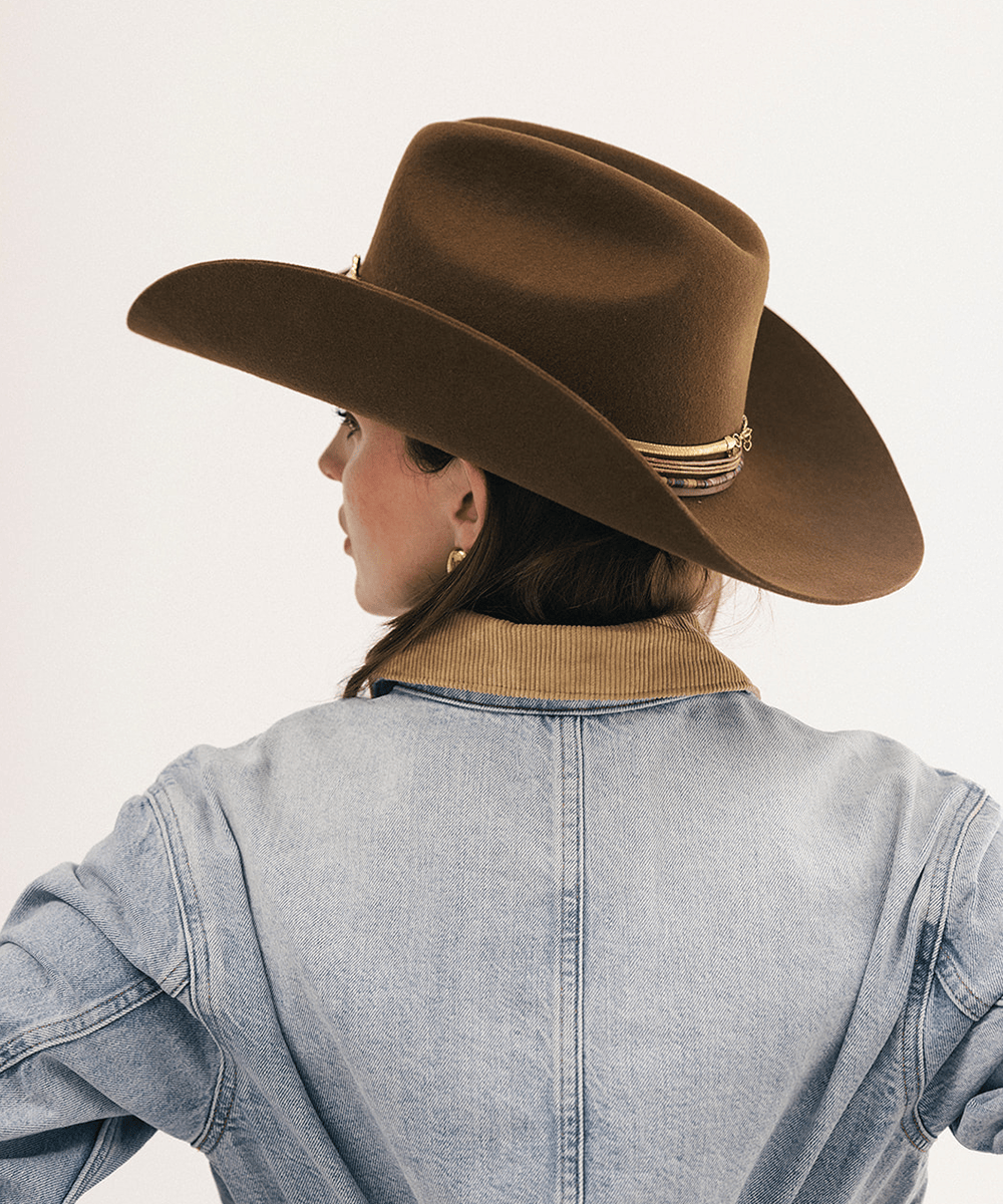 A woman shown from the back wearing a brown felt cowboy hat with a gold band and a light blue denim jacket against a plain white background #color_coffee