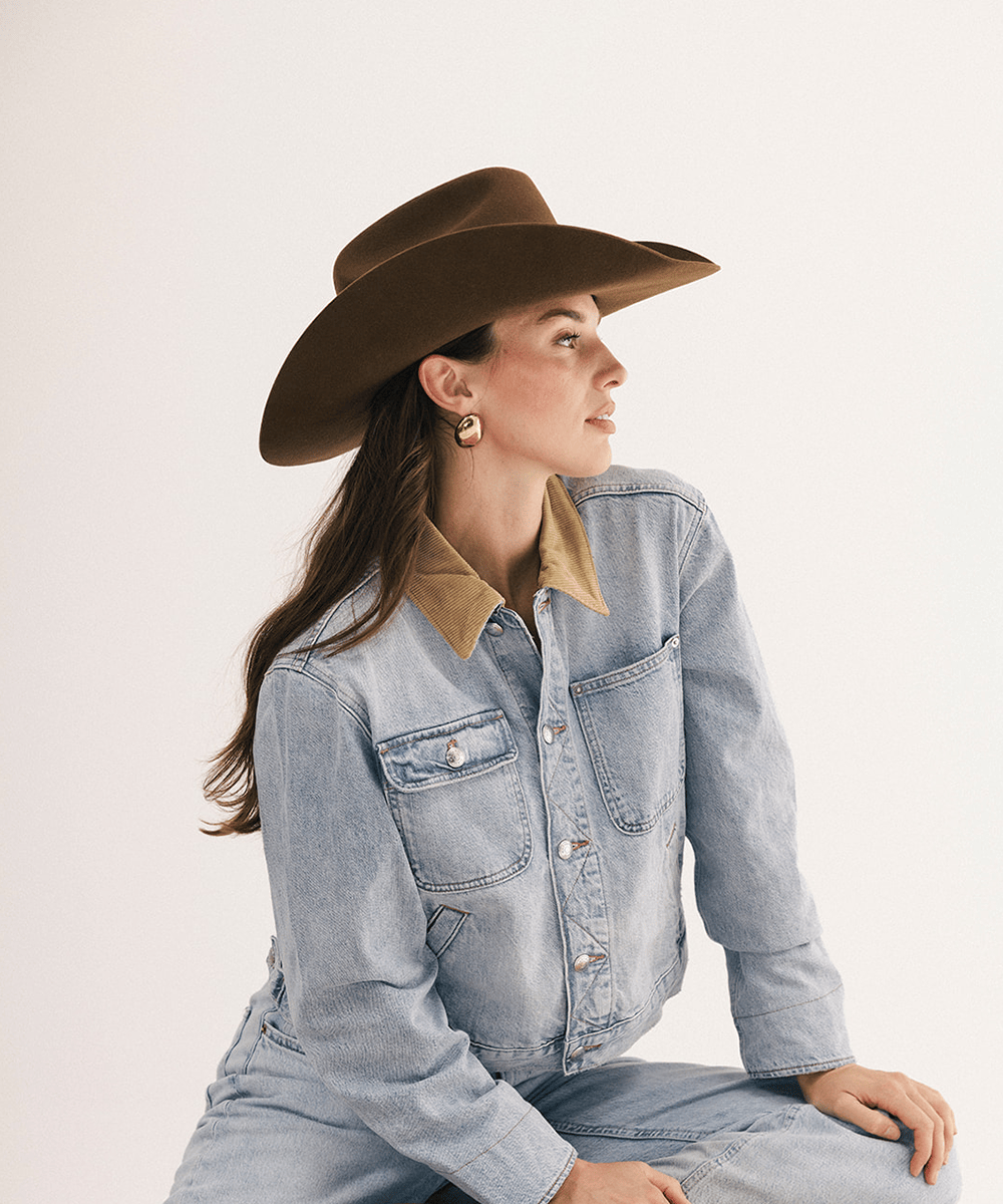 A woman wearing a brown wide-brim hat, a light-wash denim jacket with a tan collar, and matching light-wash denim jeans, posed against a plain off-white studio background #color_coffee