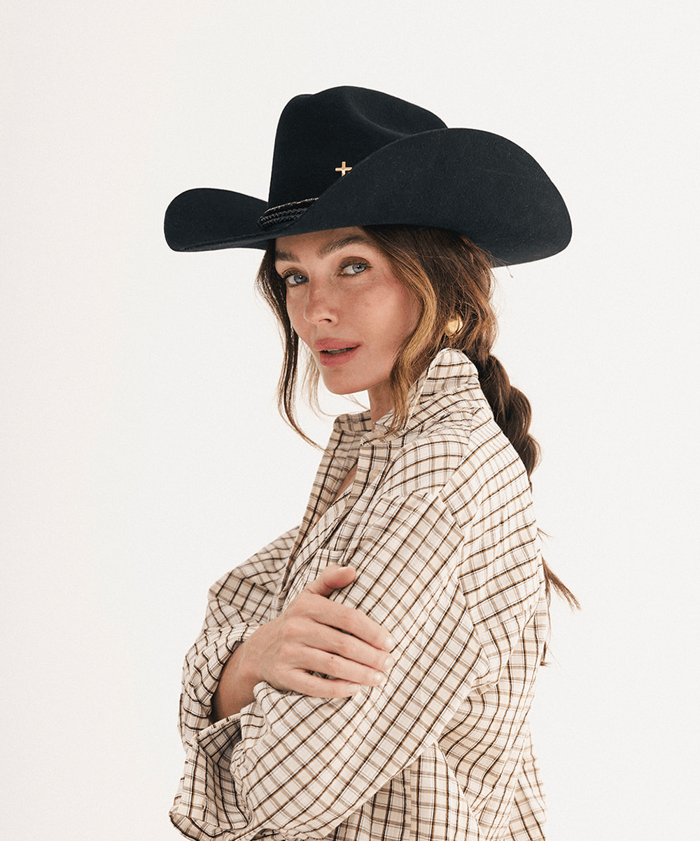 A woman wearing a black cowboy hat with a small gold cross emblem and a beige-and-brown plaid button-up shirt, photographed against a plain light background #color_black