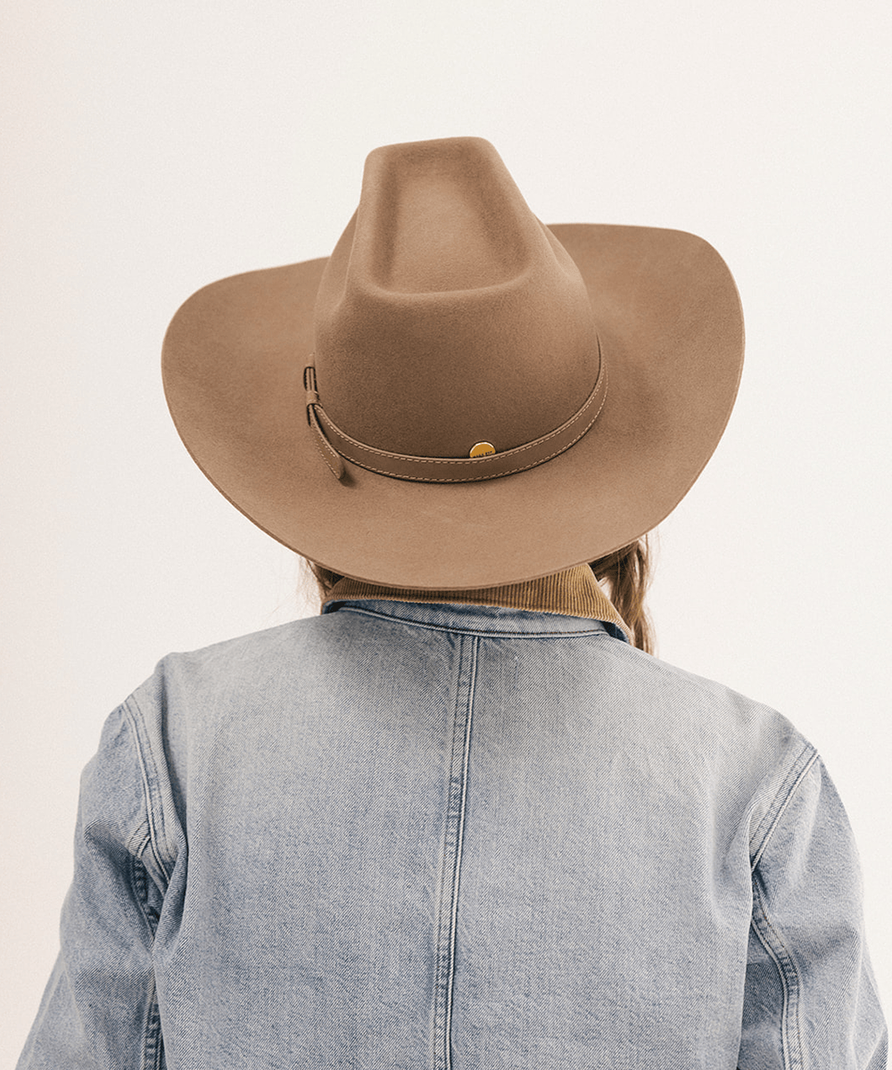 A person viewed from behind wearing a wide-brim tan cowboy hat with a matching band and a light blue denim jacket against a plain off-white background #color_brown