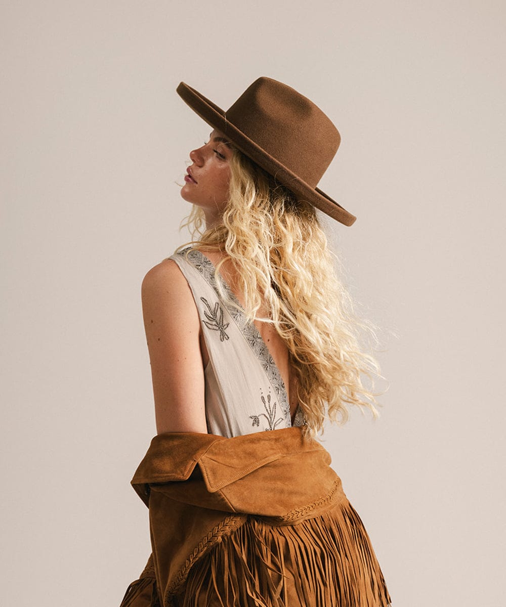 Woman wearing a chocolate colored pencil brim felt fedora hat and fringed jacket, facing sideways against a plain background #color_chocolate