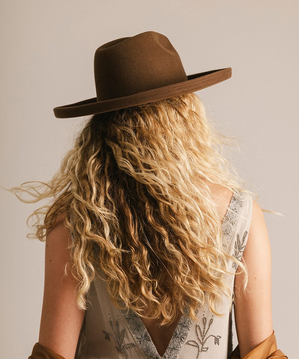 Woman wearing a chocolate colored pencil brim felt fedora hat facing behind against a plain background #color_chocolate