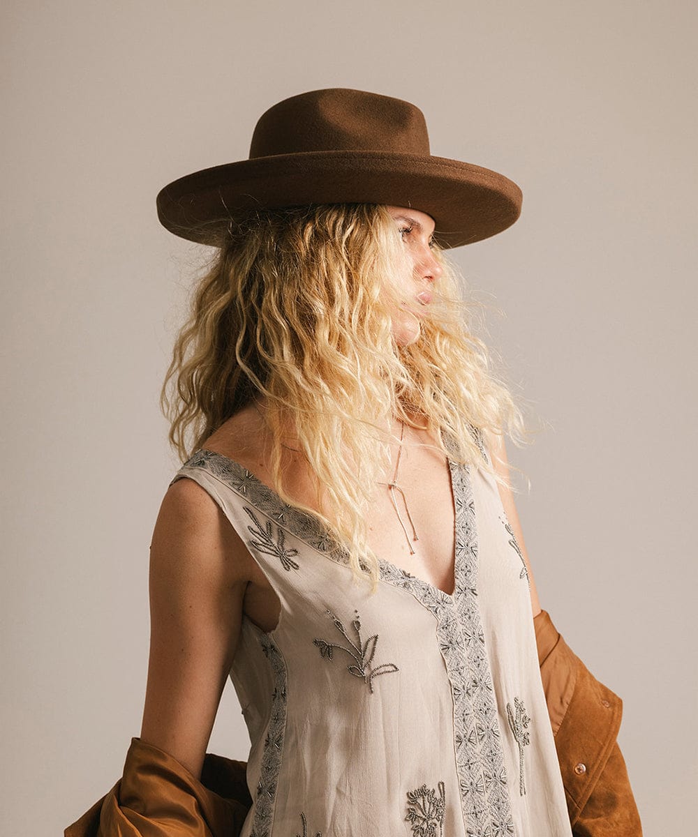 Woman wearing a chocolate colored pencil brim felt fedora hat and a white embroidered dress, looking sideways against a plain background #color_chocolate